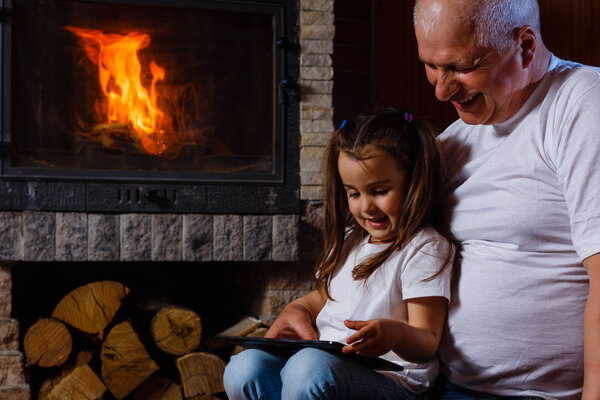 Grandfather and granddaughter having fun using digital tablet sitting at fireplace