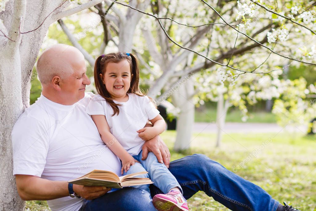 Happy grandfather with his granddaughter reading sitting in blooming garden