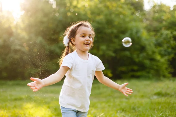 Cute curly girl playing with soap bubbles in green garden. Happy childhood concept.