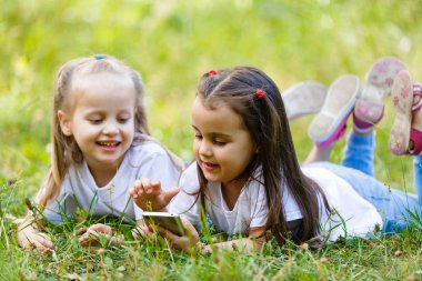 Parktaki çimlerin üzerinde yatan iki küçük gülümseyen kız. Cep telefonunda oynat