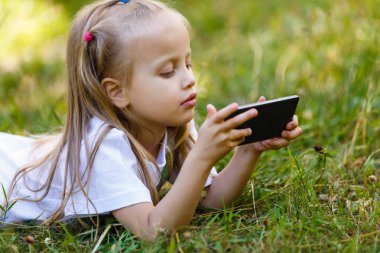 Güzellik ve moda kavramı, küçük bir kız park, yeşil çim üzerinde yalan smartphone ile portre portre