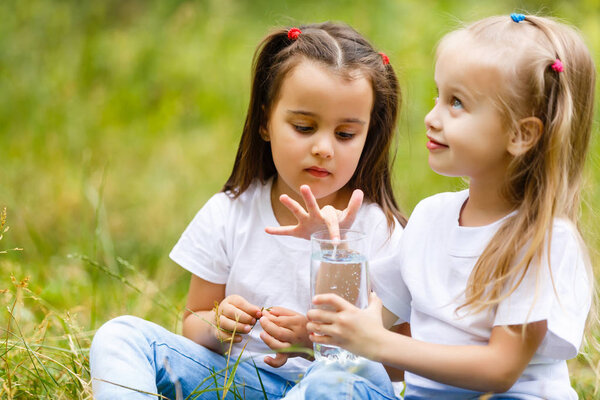 Two little girls drinking glass of water in green park. Concept of purity, ecological and biological product, love for nature