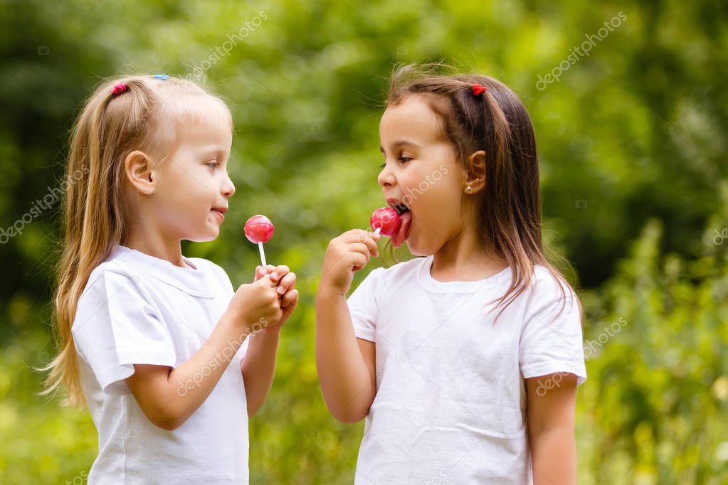 Two cute little sisters eating lollipops in green in summer park