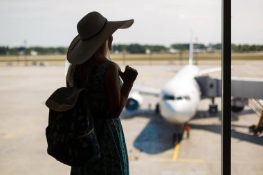 Tatil başlıyor. Modern Havaalanı terminal uçuşu için bekleyen sırt çantası ile yaz elbiseli Genç kadın bina, gündoğumu veya günbatımı, uçaklar penceresinde izlerken arkadan görünüm
