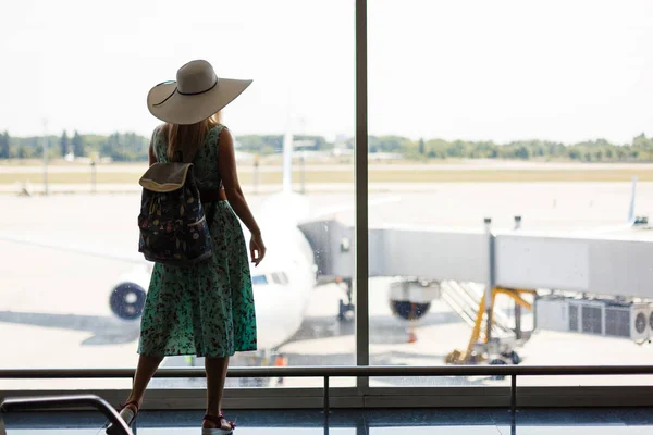假期开始了 年轻妇女在夏天礼服与背包等待飞行在现代机场终端大厦 看飞机在窗口在日出或日落 后面看法 — 图库照片
