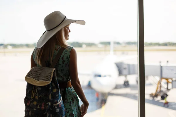假期开始了 年轻妇女在夏天礼服与背包等待飞行在现代机场终端大厦 看飞机在窗口在日出或日落 后面看法 — 图库照片