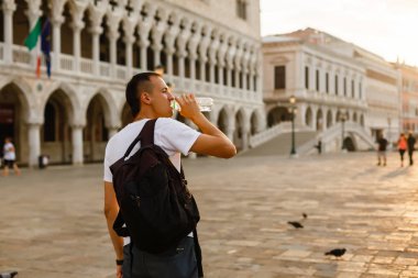 Genç yakışıklı adam içme suyu Venedik'te San Marco Meydanı'nda bir şişe beyaz t-shirt giyen şafakta, İtalya,