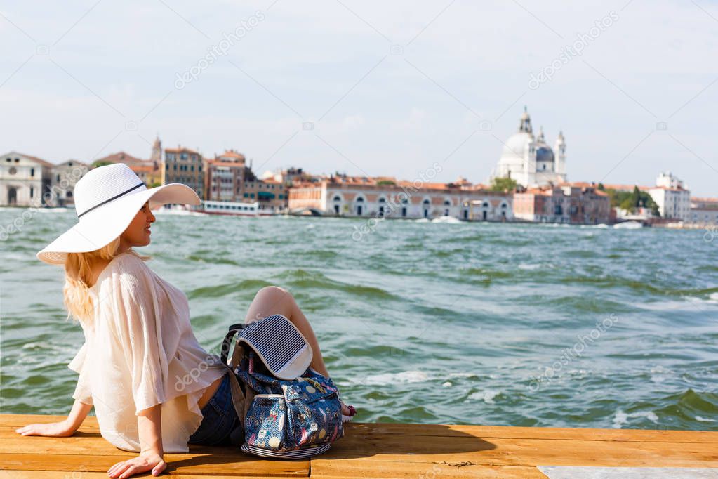 Young female traveler sitting on the pier and enjoying beautiful view on venetian chanal with gondolas floating in Venice