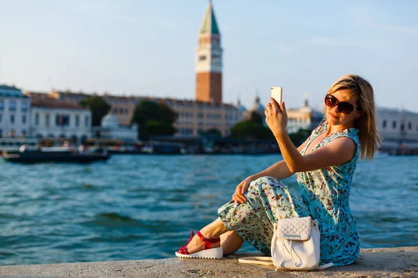 Woman sits near a canal and admires gondolas and looking into a smartphone