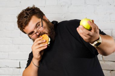 Açgözlülükle hamburger yeme ve yeşil elma beyaz zemin üzerine başka bir elinde tutan sakallı adam heyecanlı