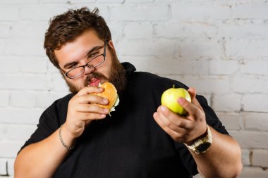 Açgözlülükle hamburger yeme ve yeşil elma beyaz zemin üzerine başka bir elinde tutan sakallı adam heyecanlı