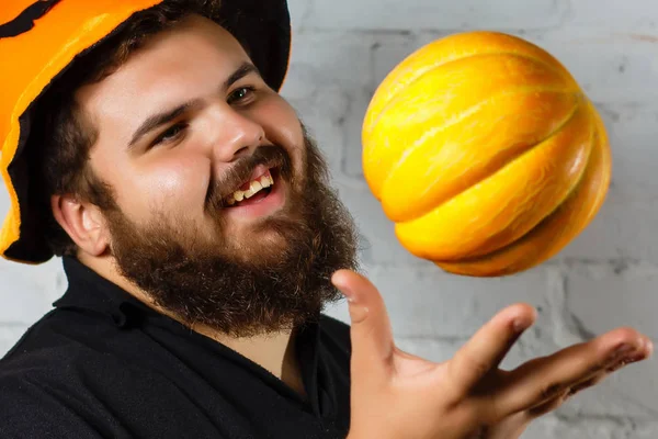 Halloween hipster with beard smiling in wizard hat. Man with pumpkin on white brick wall. Trick or treat. Holiday celebration concept.