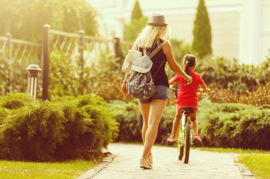 Güzel ve mutlu genç bir anne tatlı kızına bisiklet sürmeyi öğretiyor. Hem gülümsüyor hem de birbirlerine bakıyorlardı. Arka planda yaz parkı