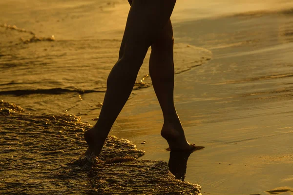 Gün batımında kumlu plajda okyanus dalgaları üzerinde yürüyen bacaklar siluetinin yakın görünümü