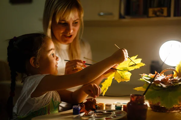 little girl with mother drawing with paint autumn leaves at home