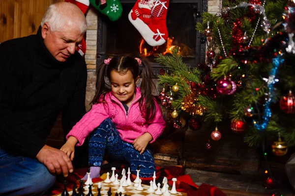 Grandfather teaching granddaughter to play chess near decorated fireplace and Christmas tree in cottage