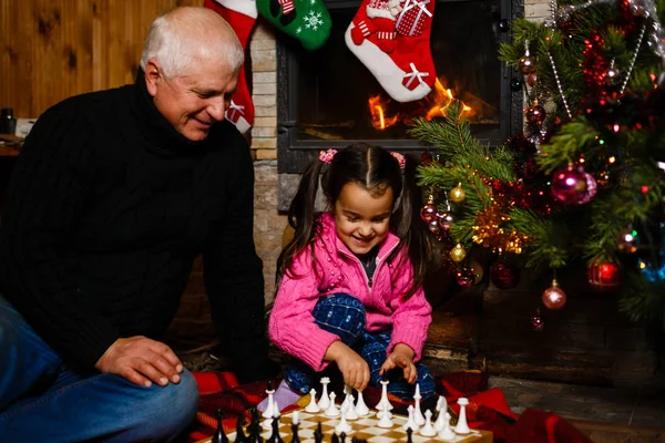 Grandfather teaching granddaughter to play chess near decorated fireplace and Christmas tree in cottage