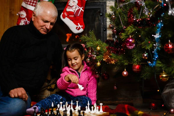 Grandfather teaching granddaughter to play chess near decorated fireplace and Christmas tree in cottage