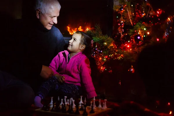 Grandfather teaching granddaughter to play chess near decorated fireplace and Christmas tree in cottage