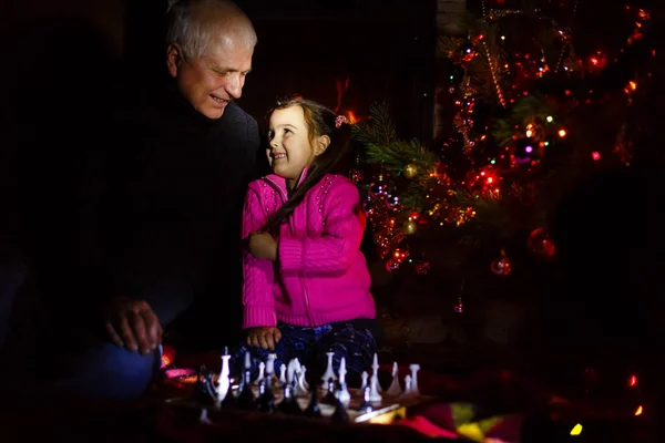 Grandfather teaching granddaughter to play chess near decorated fireplace and Christmas tree in cottage