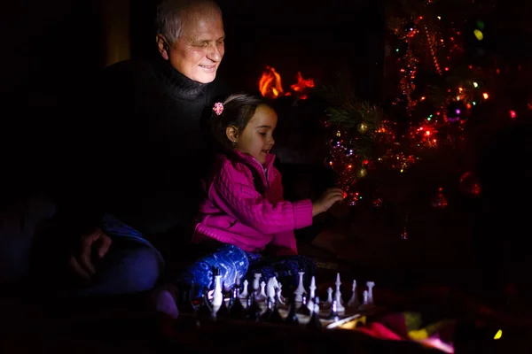 Grandfather teaching granddaughter to play chess near decorated fireplace and Christmas tree in cottage