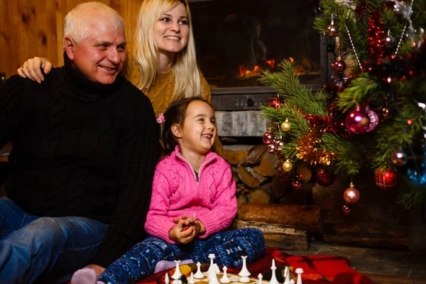 Grandfather and mother teaching granddaughter to play chess near decorated fireplace and Christmas tree in cottage