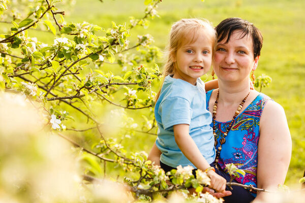 Happy mother holding preschool daughter and standing beside blooming tree 