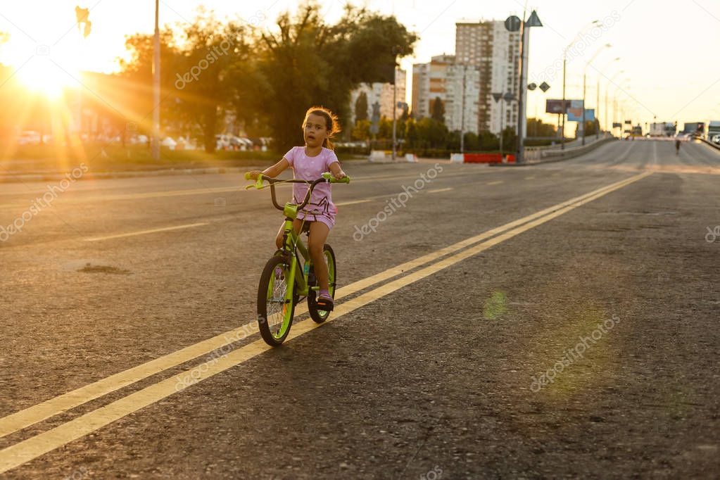 Little brunette girl riding bicycle on road at sunset