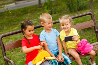 Üç mutlu çocuk parkta bankta smartphone ile oynamak 