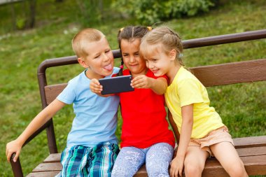 Üç mutlu çocuk parkta bankta smartphone ile oynamak 