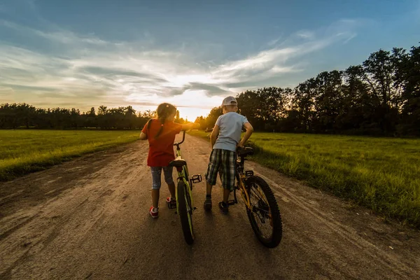 children riding one bike together on sunny summer evening. sitting on bicycle rack. Family of two people enjoying traveling in scenic field over sunset sky background.