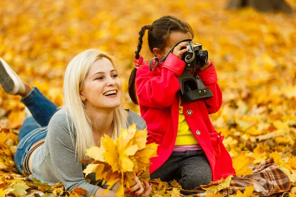 mother and daughter having fun with retro camera while resting on autumn orange leaves outdoor