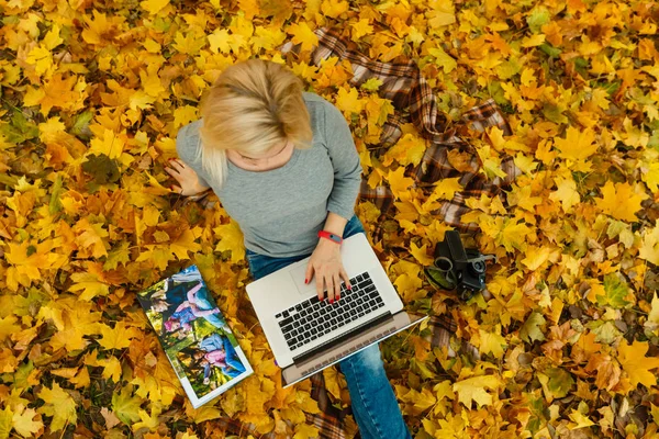 woman using laptop and family album while sitting on autumn orange leaves at park