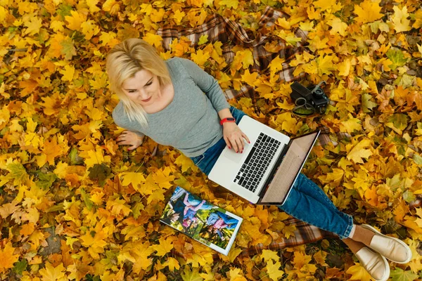 woman using laptop and family album while sitting on autumn orange leaves at park