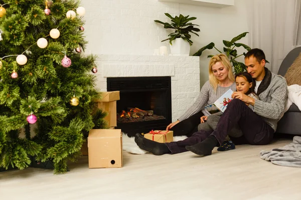 Family in with book sitting on floor near Christmas tree at living room