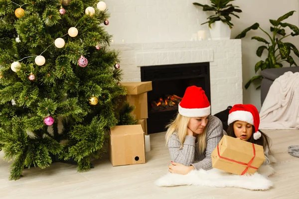 現在リビングのクリスマス ツリーの近くの床に横たわっていると赤いサンタ帽子の娘を持つ母 — ストック写真