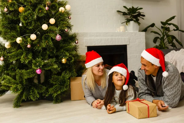 現在リビングのクリスマス ツリーの近くの床に横たわっていると赤いサンタ帽子の家族 — ストック写真