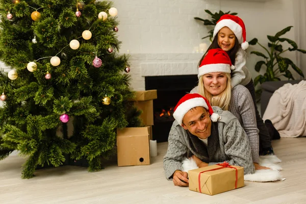 現在リビングのクリスマス ツリーの近くの床に横たわっていると赤いサンタ帽子の家族 — ストック写真