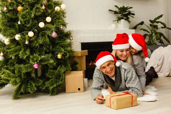 現在リビングのクリスマス ツリーの近くの床に横たわっていると赤いサンタ帽子の家族 — ストック写真