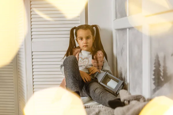 pretty little girl in stylish clothes with christmas vintage lantern sitting on window sill