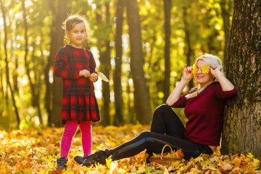 Genç anne ve kızı bir sonbahar parkında yaprak topluyor.