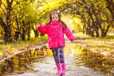 Pembe ceketli ve lastik çizmeli çok tatlı bir kız yağmurlu bir günde su birikintisinin üzerinden atlıyor.