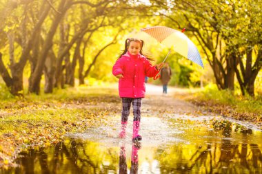 Yağmurda şemsiyesi olan mutlu bir kız çocuğu su birikintilerinde koşuyor.