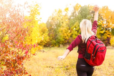 Sırt çantalı genç kadın elini kaldırıyor ve sonbahar parkının keyfini çıkarıyor.