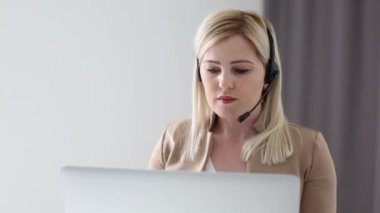 Kendine güvenen kadın ofis çalışanı kulaklık takıyor ve dizüstü bilgisayara bakıyor. Genç iş kadını müşteri hizmetleri müdürü uzaktan iletişim kuran webcam ile konuşuyor..