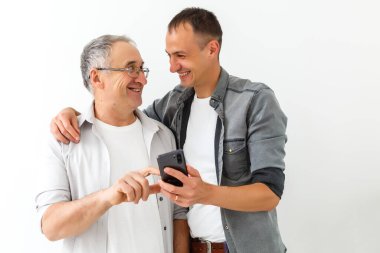 Mutlu iki nesil erkekler aile babası genç yetişkin oğlunu kucaklıyor akıllı telefon bağlantısı kurarak eğleniyor mobil uygulamalar kullanarak komik sosyal medya videoları izliyorlar.