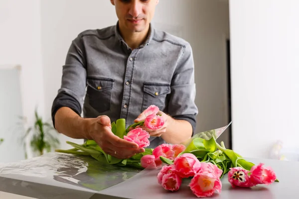Man florist makes red tulip bouquet and wrapping in pack on wooden table. Flowers