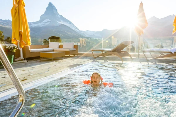 Little girl playing in outdoor swimming pool of luxury spa alpine resort in Alps mountains, Austria. Winter and snow vacation with kids. Hot tub outdoors with mountain view. Children play and swim.