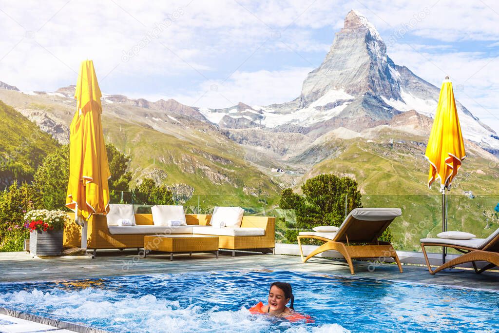 Kids play in outdoor infinity swimming pool of luxury spa alpine resort at sunset in Alps mountains. Spring or summer vacation for family with children.