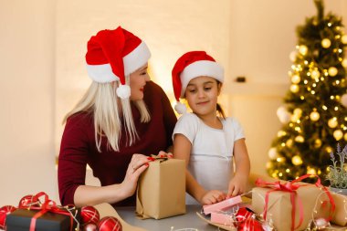 Tatiller, kutlamalar, aile ve insanlar konsepti. Mutlu anne ve küçük kız Noel Baba şapkalı, hediye kutusu oturma odasında ve Noel ağacı arka planında.